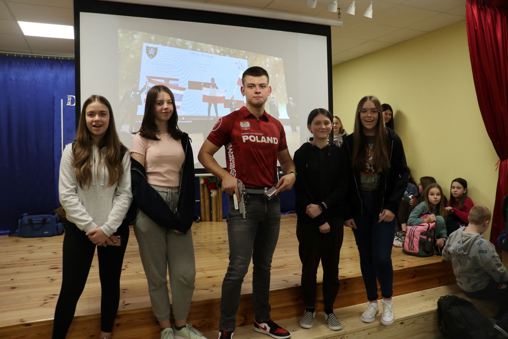 Uczennice Szkoły Podstawowej wraz z gościem Dawidem Chojnowskim stojący przed ekranem na którym odtwarzany jest film.