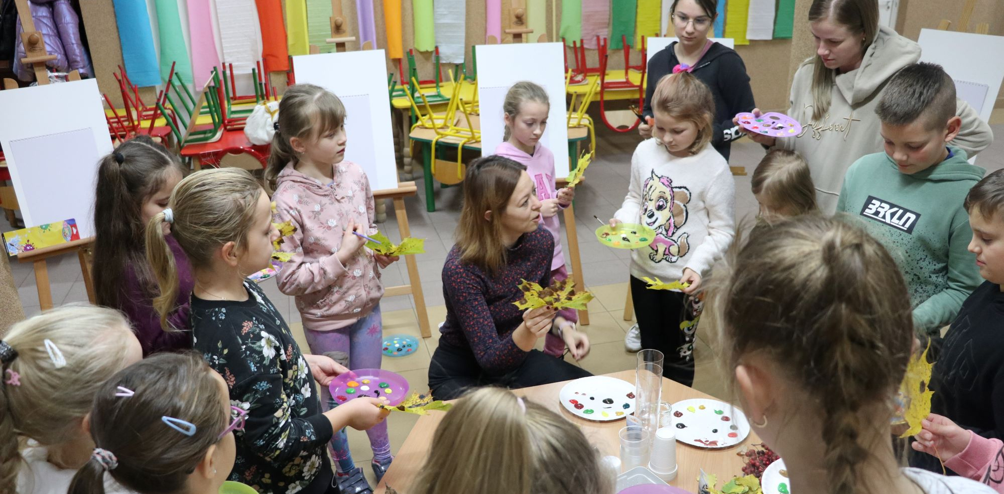 Grupa dzieci stoi wokół stolika przy którym prowadząca zajęcia Pani Anna Dziedziul prezentuje jesienny liść. Dzieci trzymaja w rękach palety z farbami.